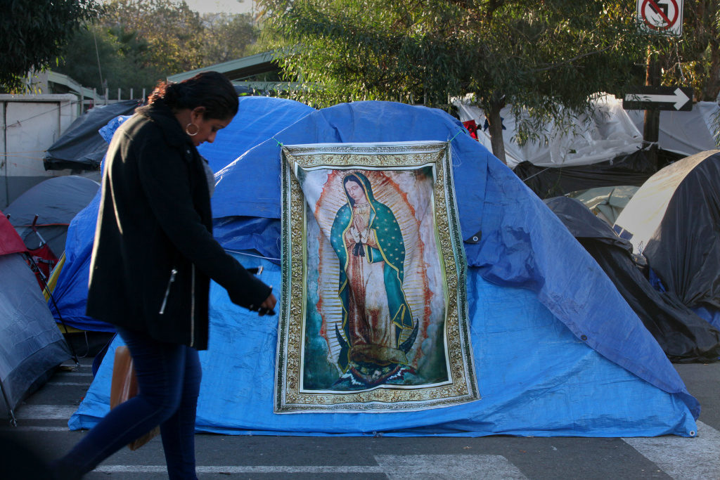 At the Benito Juarez Sports Complex near downtown Tijuana,  a woman walks past a tapestry of the Virgen de Guadalupe in the middle of the migrant tents on Dec. 16, 2018.